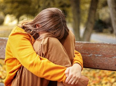 Lonely upset heartbroken teenager girl crying sitting on bench outdoors hiding her face in knees. Problems of teenage age. Autumn seasonal melamcholy.