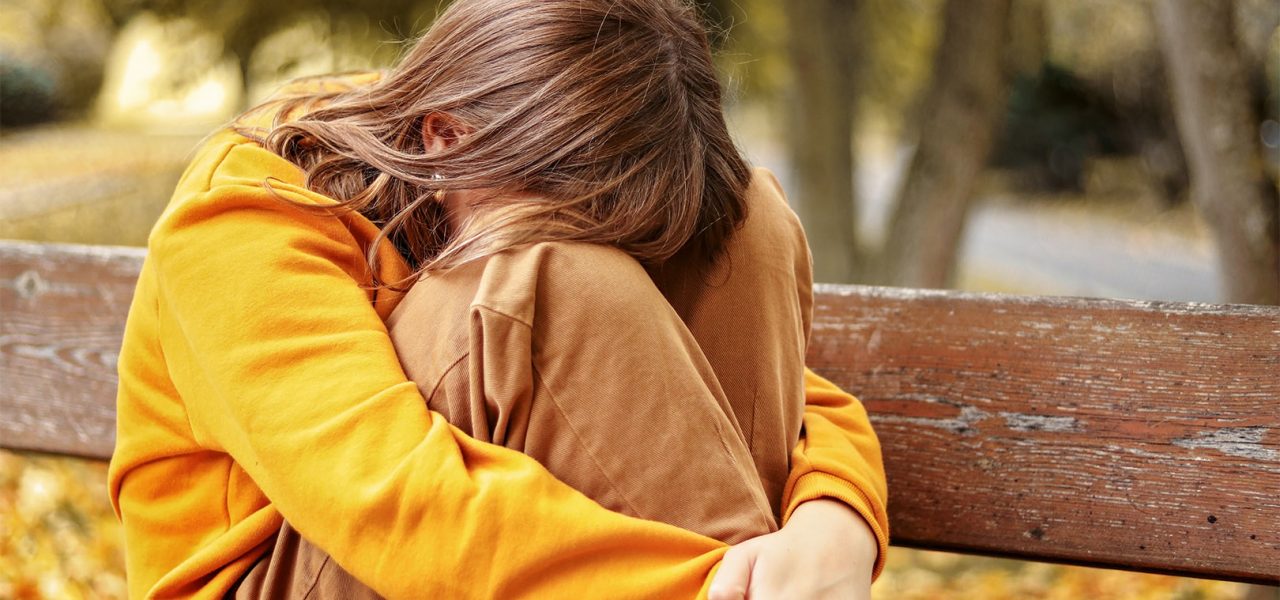 Lonely upset heartbroken teenager girl crying sitting on bench outdoors hiding her face in knees. Problems of teenage age. Autumn seasonal melamcholy.