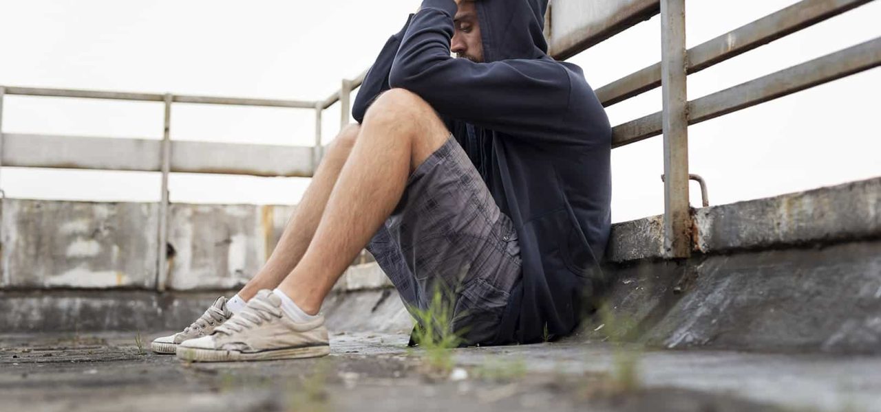 Depressed, desperate young man sitting on the building rooftop terrace, wearing hoodie, holding head in hands