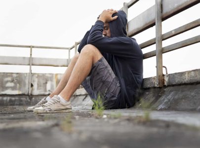 Depressed, desperate young man sitting on the building rooftop terrace, wearing hoodie, holding head in hands