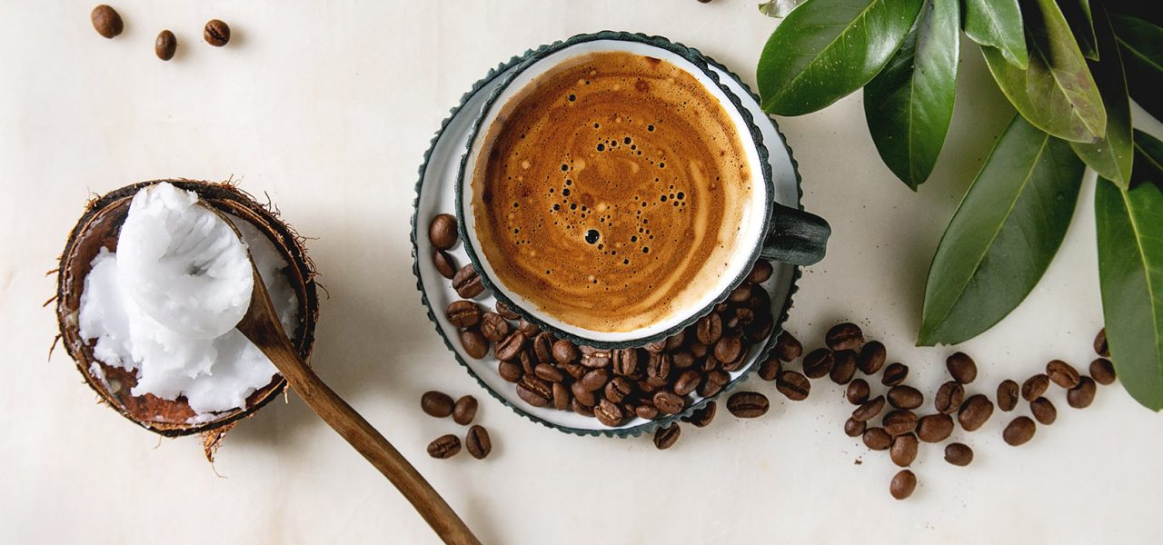 Bulletproof coffee. Keto diet coffee in blue ceramic cup with organic coconut cold press oil in wooden spoon with beans and green branch over white marble background. Flat lay, space