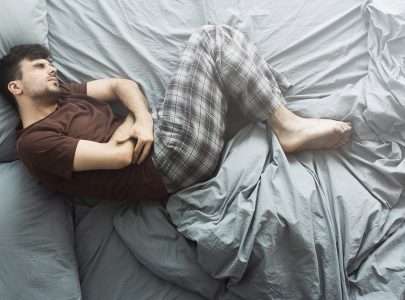 Young man suffering from stomach ache, lying in bed at home, top view. Health problems concept