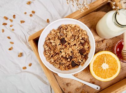 Good morning. Breakfast on white bed sheets. Muesli or granola, milk, orange  on wooden tray from above. Top view.  Flat lay. Copy space. Hotel Room Early Morning.